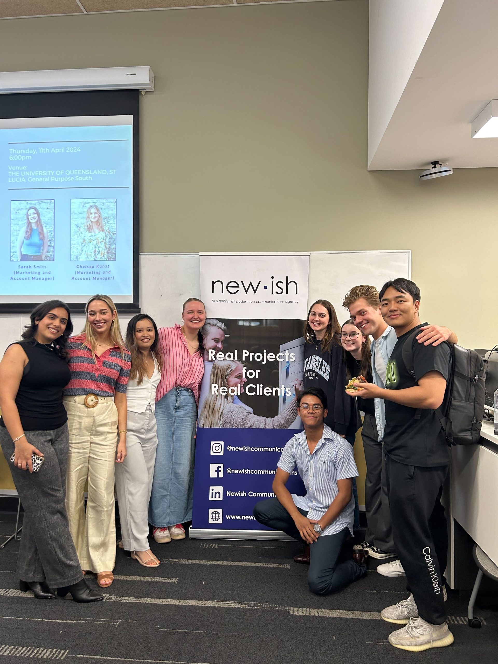 A group of people stand around the Newish sign stand.