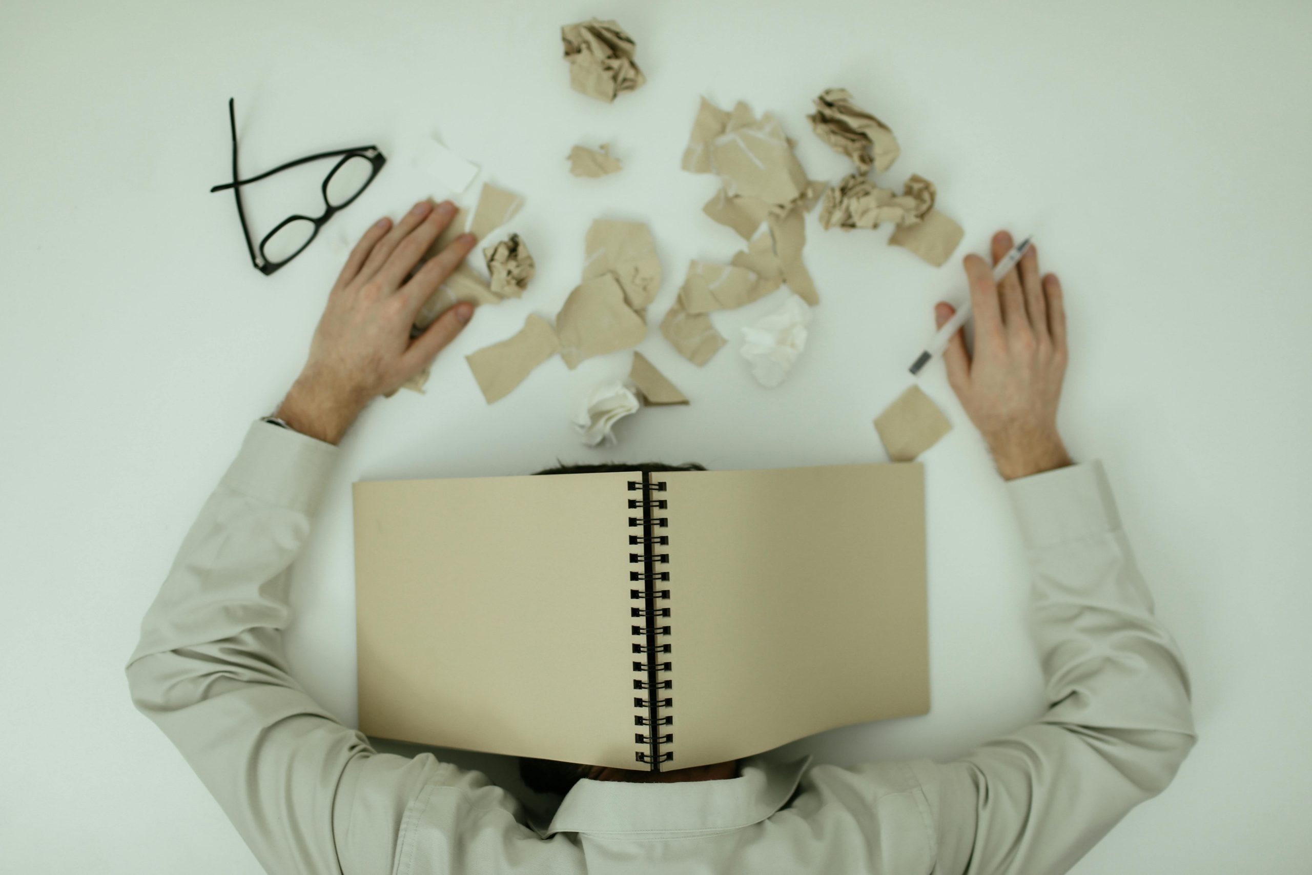 A person lies face down onto a table, their head covered by an open notebook. Scribbled-on notes and a pair of glasses are laid out messily around them. They are tired and experiencing burnout.